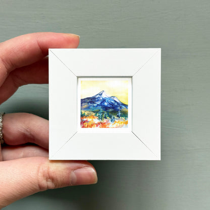 A hand is holding a small white square frame containing a painting of a mountain landscape.