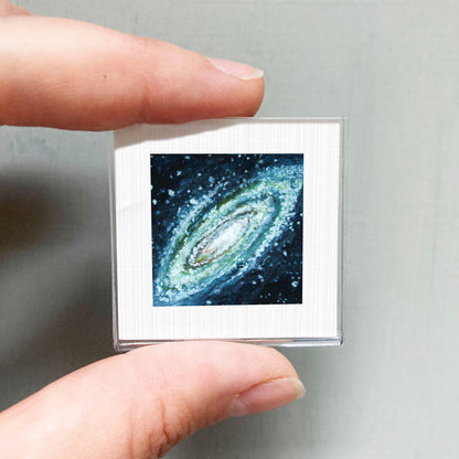 A person's hand is holding a small square frame containing a photograph of a spiral galaxy.