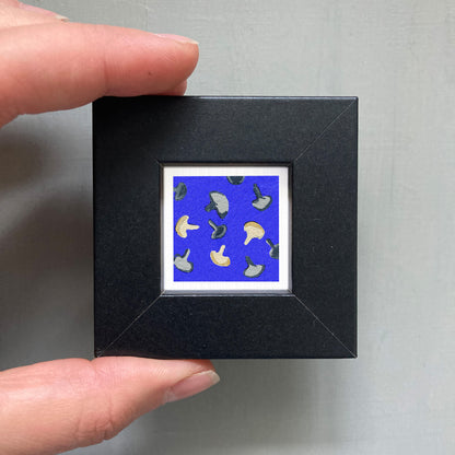 A hand is holding a small square picture frame containing a vibrant blue background with a pattern of mushrooms.