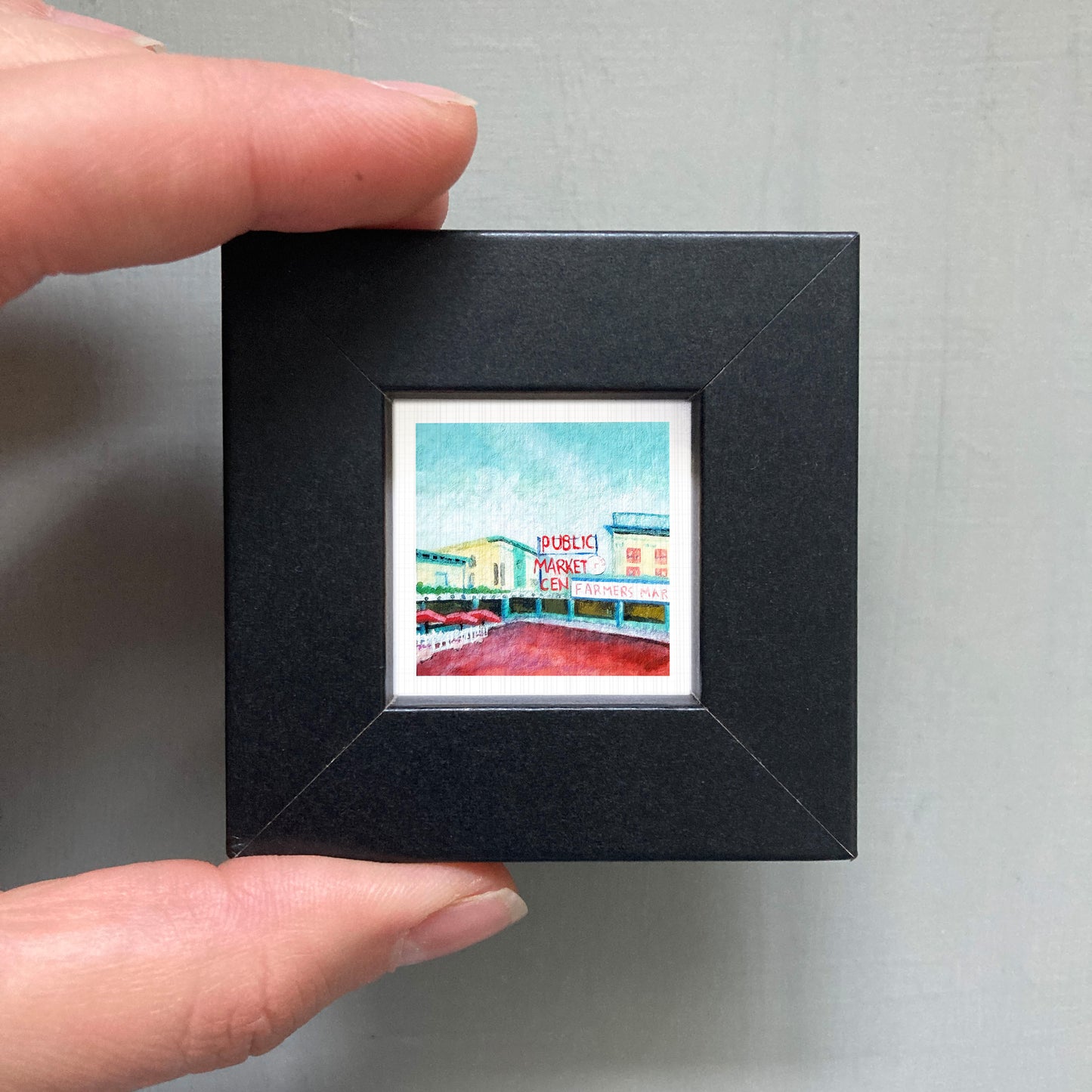 A hand is holding a small square picture frame containing a colorful painting of a building.