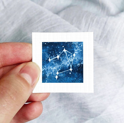 A hand is holding a small square-shaped paper with a depiction of a starry night sky on it.