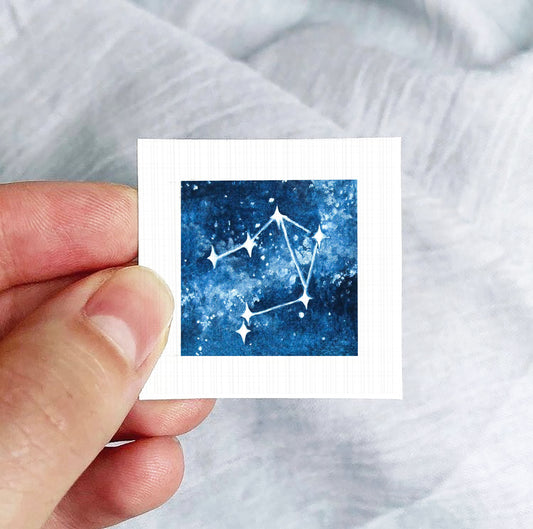 A hand is holding a small square-shaped paper with a depiction of a starry night sky on it.