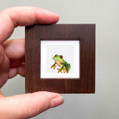 A person's hand is holding a small wooden frame containing a framed image of a green frog.