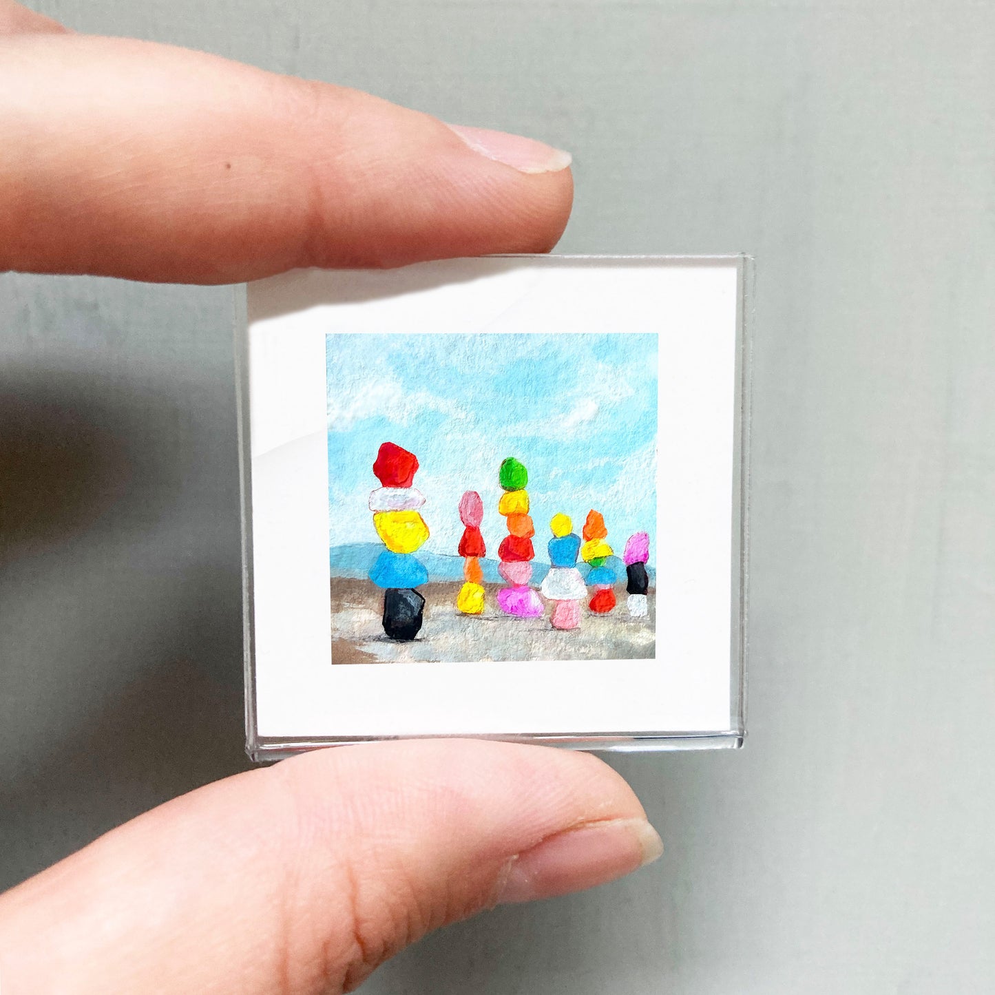A person's hand is holding a small square picture depicting a colorful scene with various objects, including a group of people and a beach landscape.