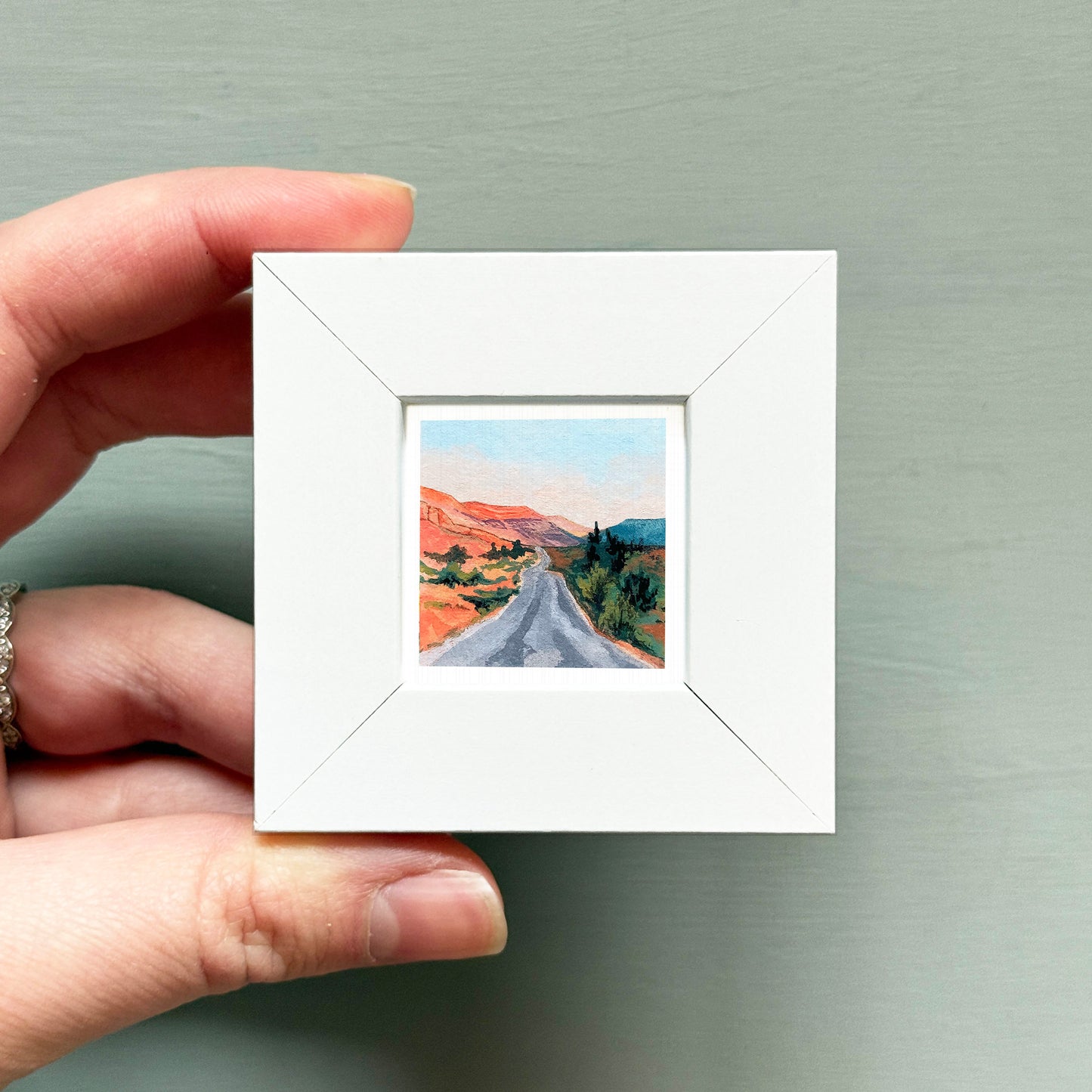 A hand is holding a small white square frame containing a painting of a winding road through a desert landscape.