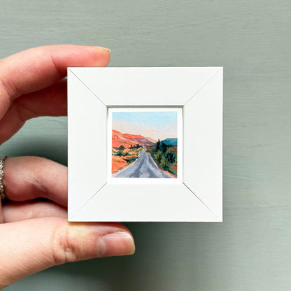 A hand is holding a small white square frame containing a painting of a winding road through a desert landscape.