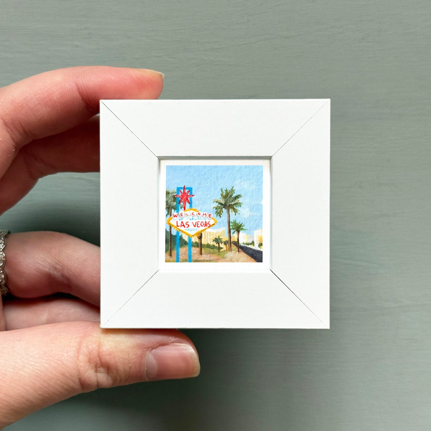 A hand is holding a small white picture frame containing a miniature painting of the Las Vegas sign.