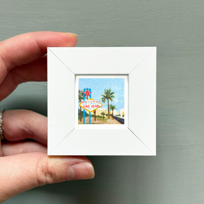 A hand is holding a small white picture frame containing a miniature painting of the Las Vegas sign.
