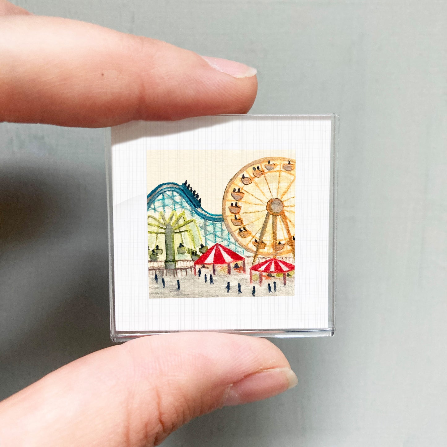 A person's hand holding a small square picture depicting an amusement park scene with a ferris wheel, roller coaster, and other rides.