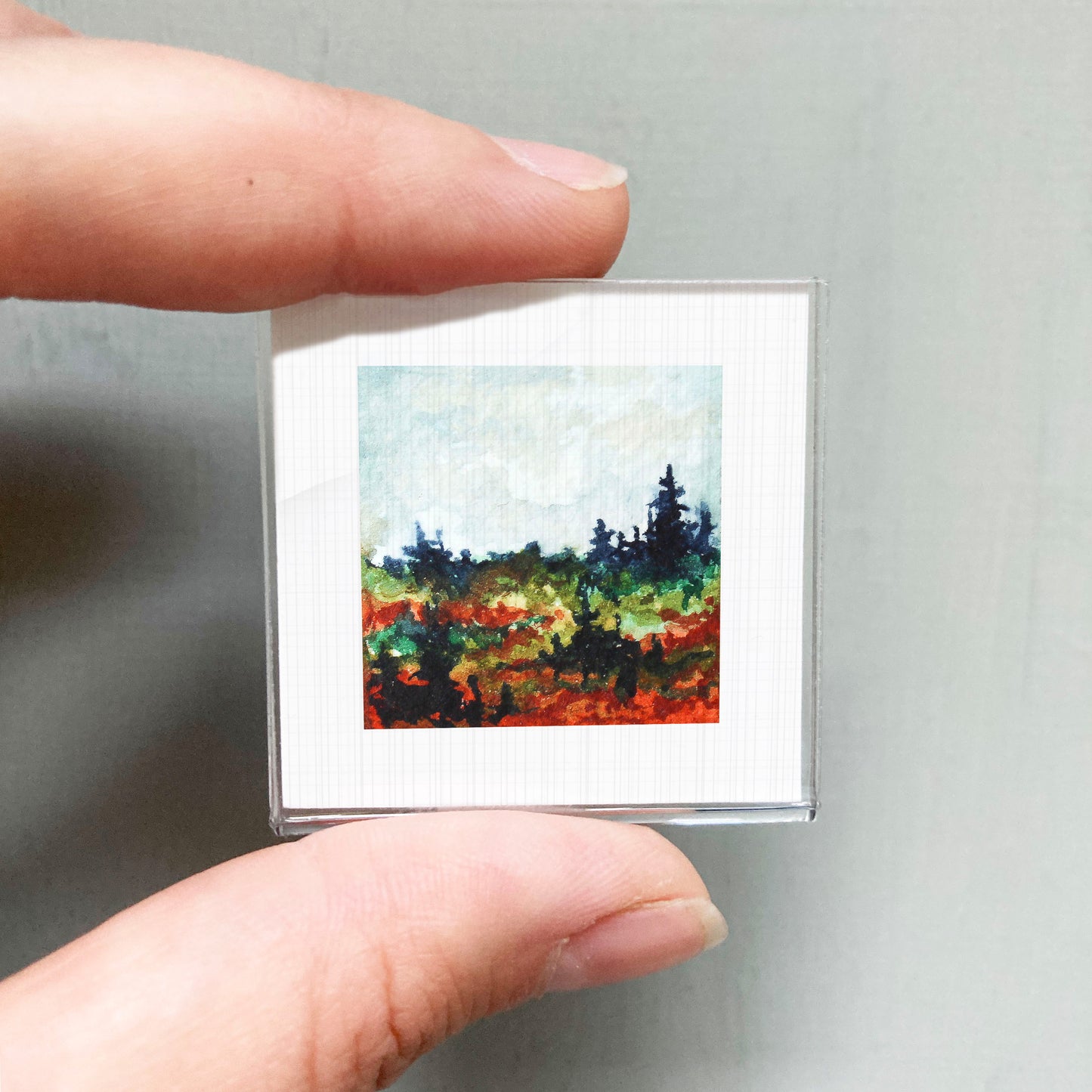A person's hand is holding a small square picture of a landscape with trees and a colorful background.