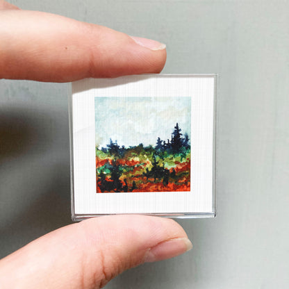 A person's hand is holding a small square picture of a landscape with trees and a colorful background.