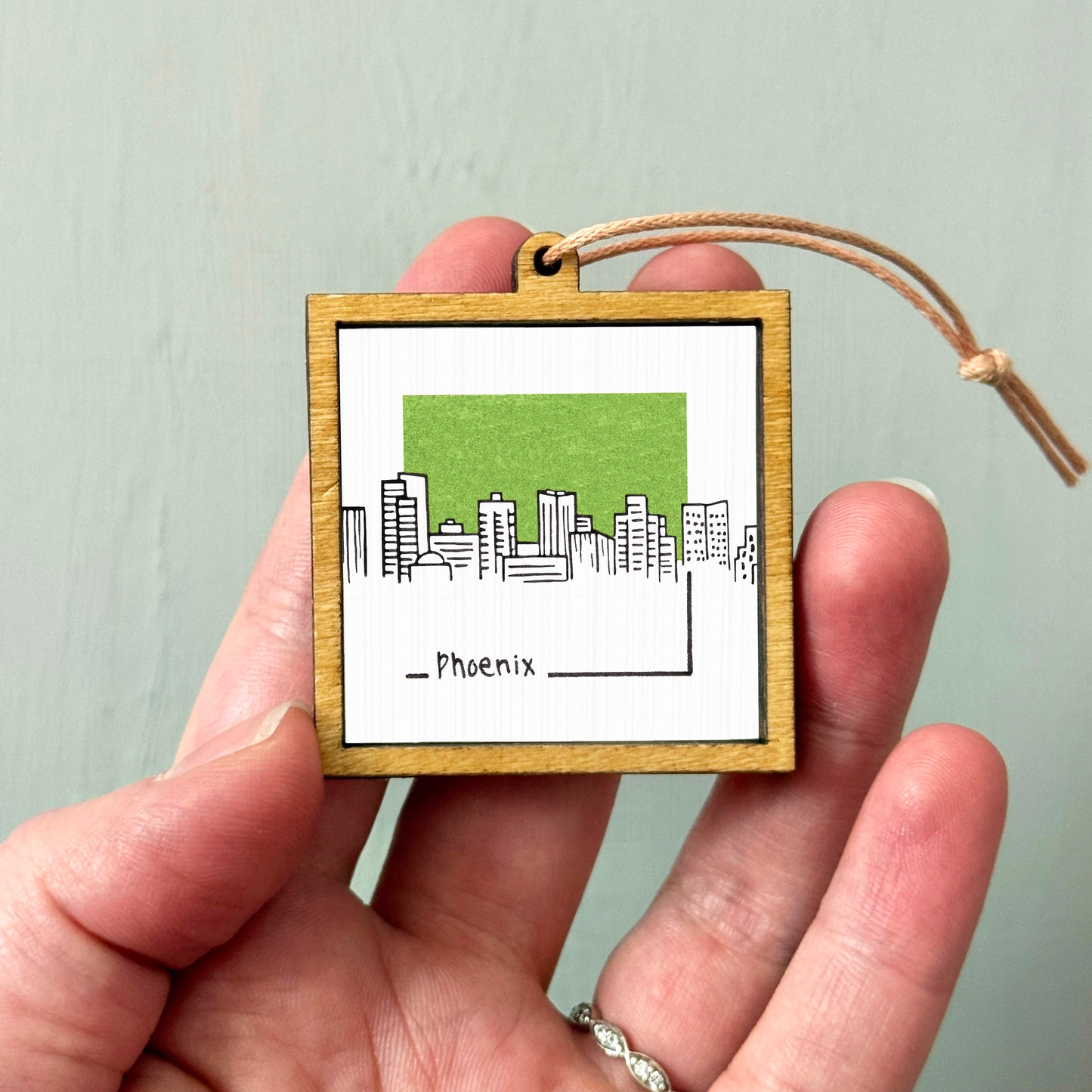 A hand holding a small wooden frame with a picture of a city skyline and the word "Phoenix" written below it.