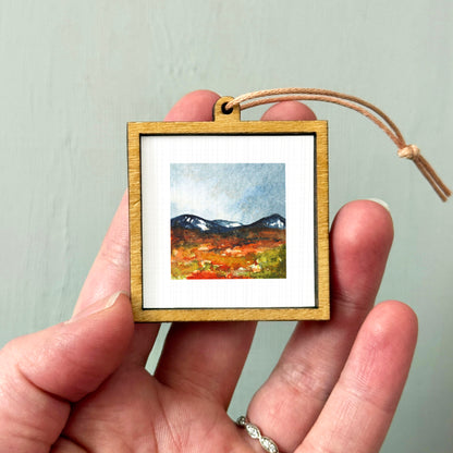 A hand holding a small wooden frame containing a painting of a mountain landscape.