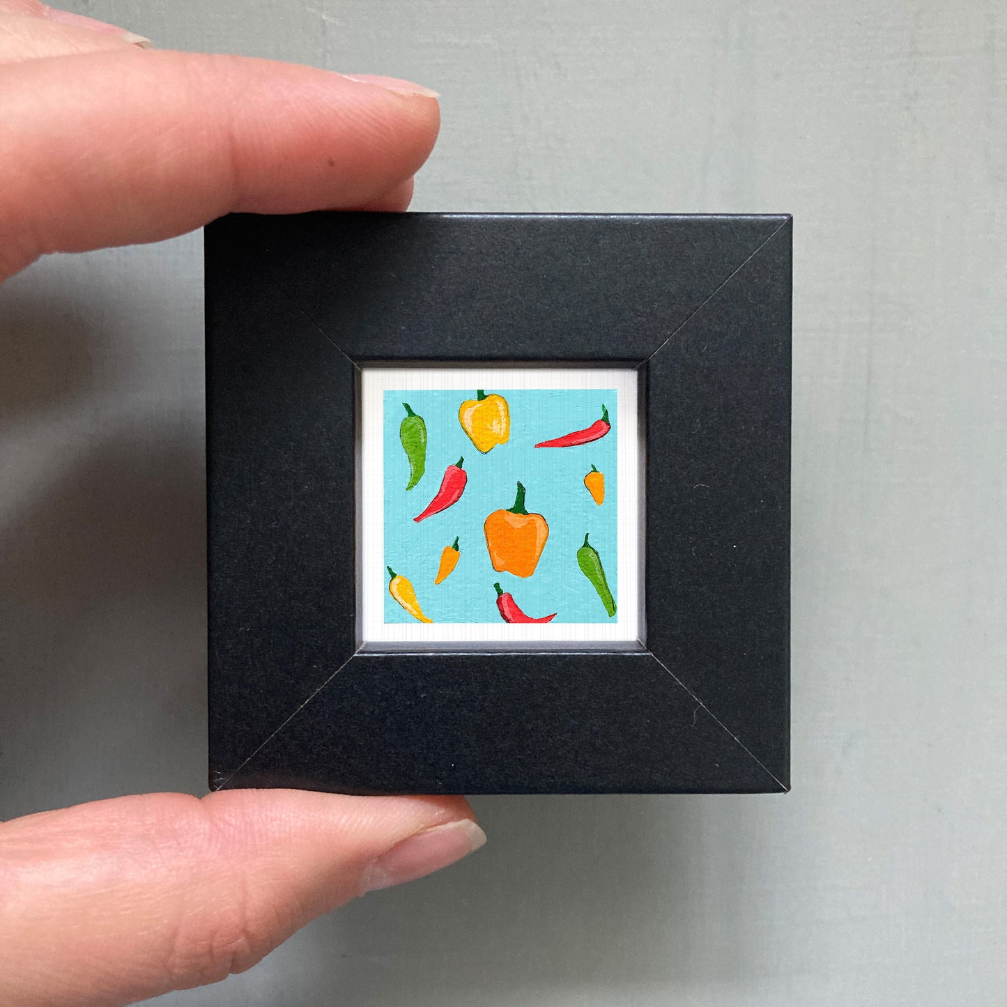 A hand is holding a small black frame containing a vibrant illustration of various peppers in different colors.