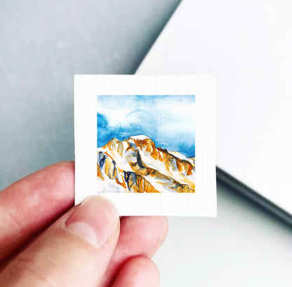 A hand is holding a small square piece of paper with a watercolor painting of a mountain landscape on it.
