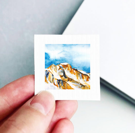 A hand is holding a small square piece of paper with a watercolor painting of a mountain landscape on it.