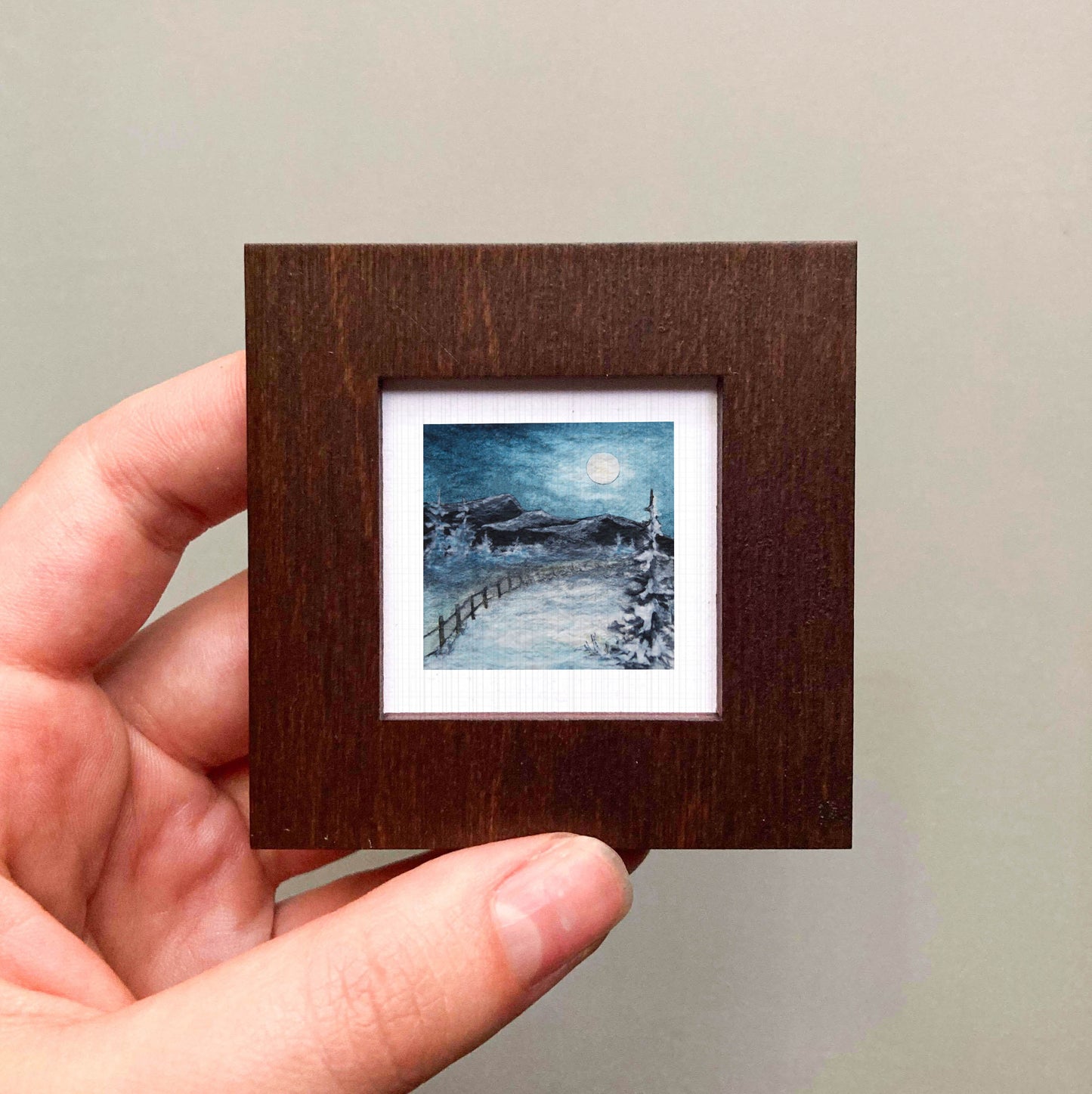 A hand is holding a small, square-shaped wooden frame containing a miniature painting of a snowy landscape with a full moon in the night sky.