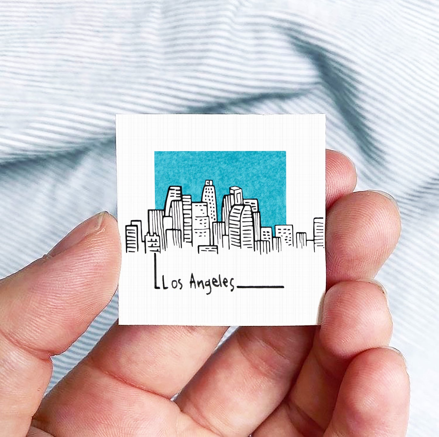 A hand holding a small square-shaped object with a drawing of a city skyline and the text "Los Angeles" written on it.