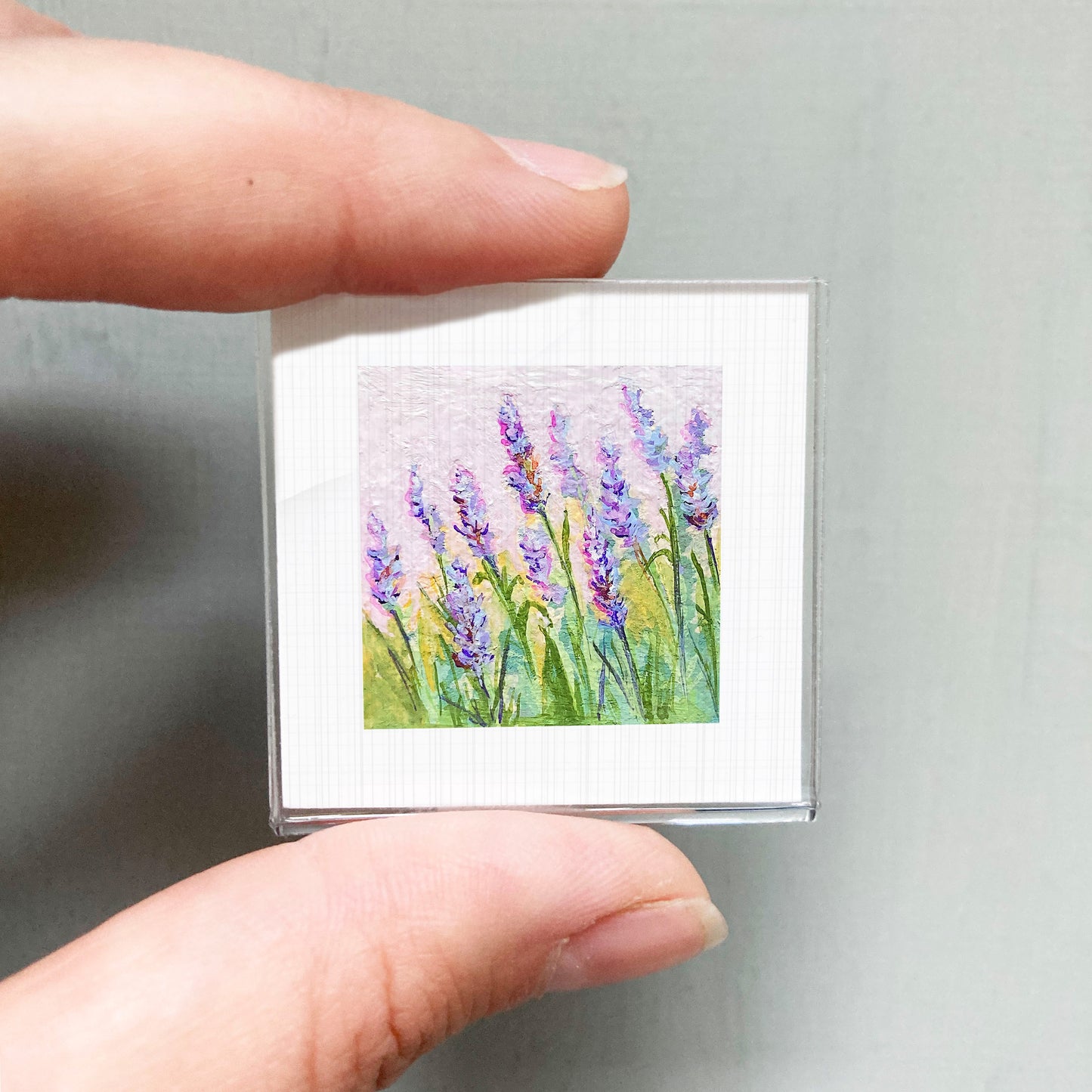 A person's hand is holding a small square picture of purple flowers against a light background.