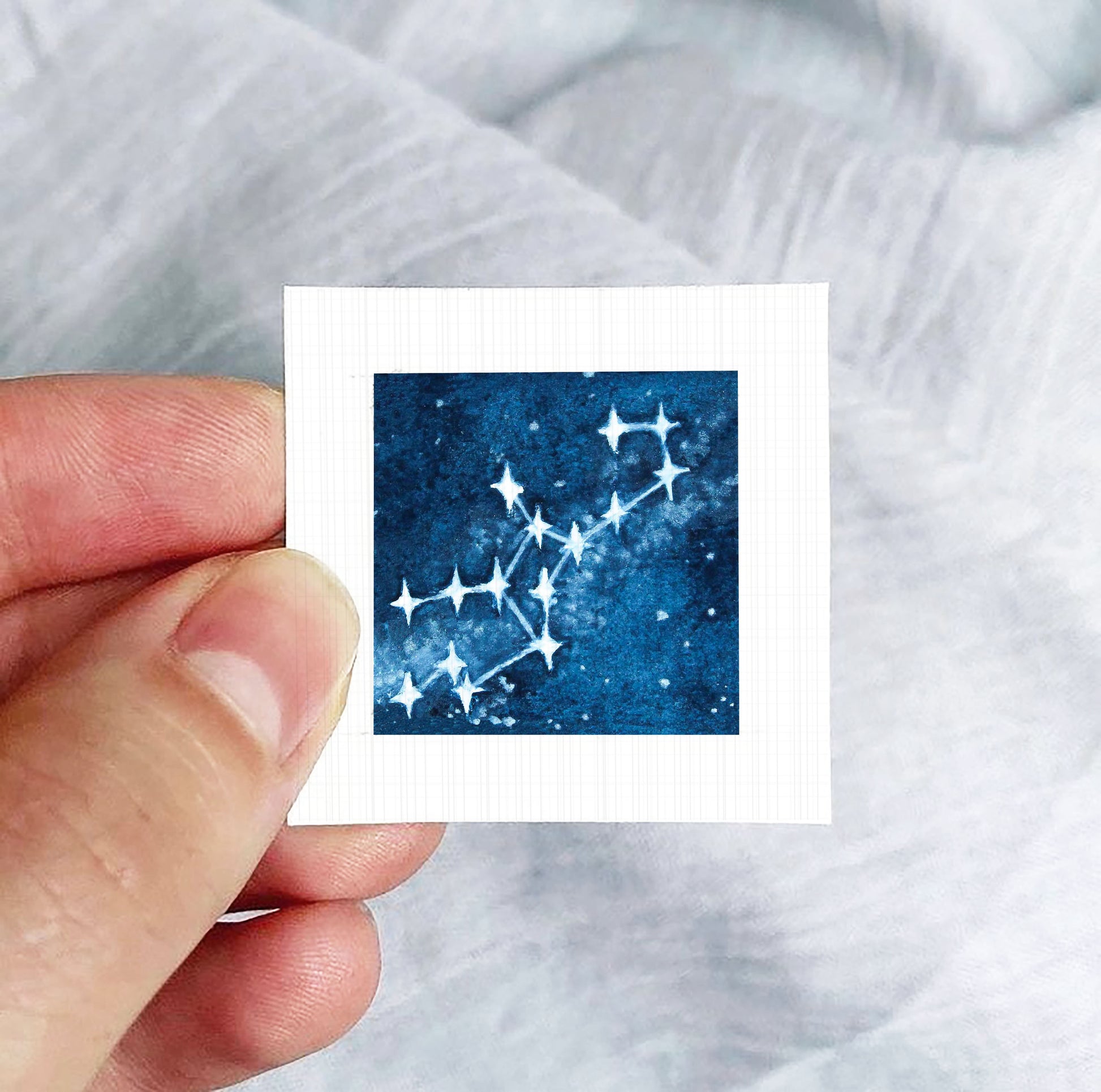 A hand is holding a small square-shaped paper with a starry night sky design on it.