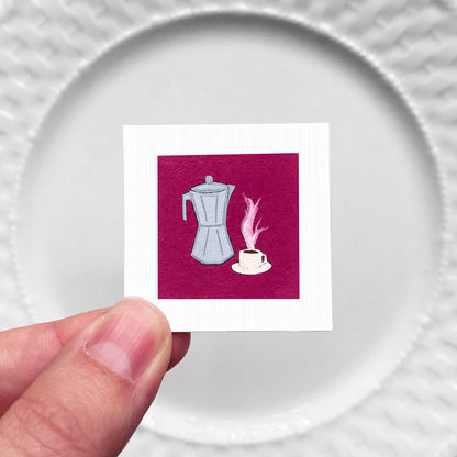 A hand is holding a small square card with an illustration of a coffee maker and a cup of coffee on it.