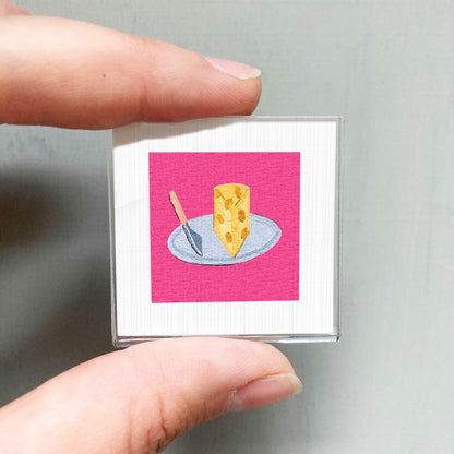 A person's hand holding a small square with a pink background and an illustration of a piece of cheese on a plate.