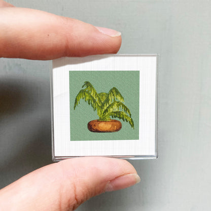 A person's hand holding a small square picture of a palm tree in a pot.