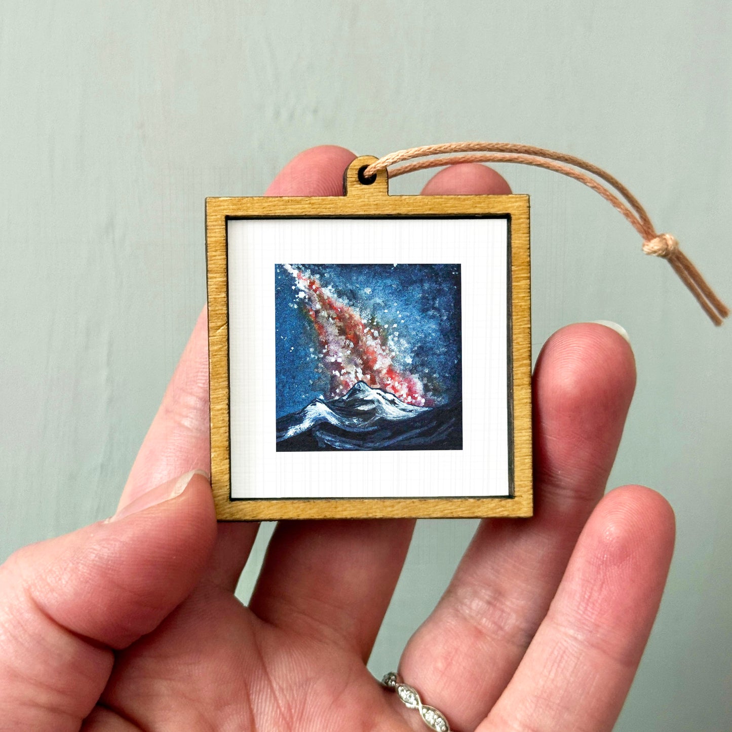 A hand holding a small wooden frame containing a painting of a blue ocean with a red and white wave.