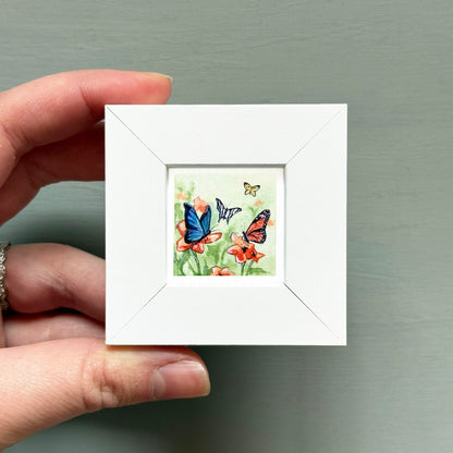 A hand is holding a small white square frame containing a painting of three butterflies on flowers.