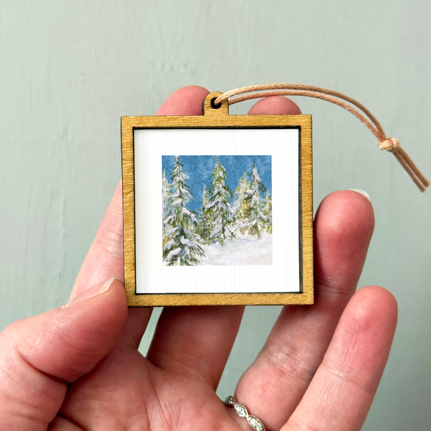 A hand holding a small wooden frame containing a picture of snow-covered pine trees.