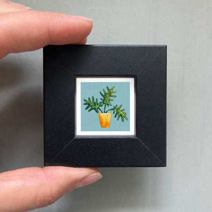 A hand is holding a small black frame containing a framed picture of a potted plant with green leaves.