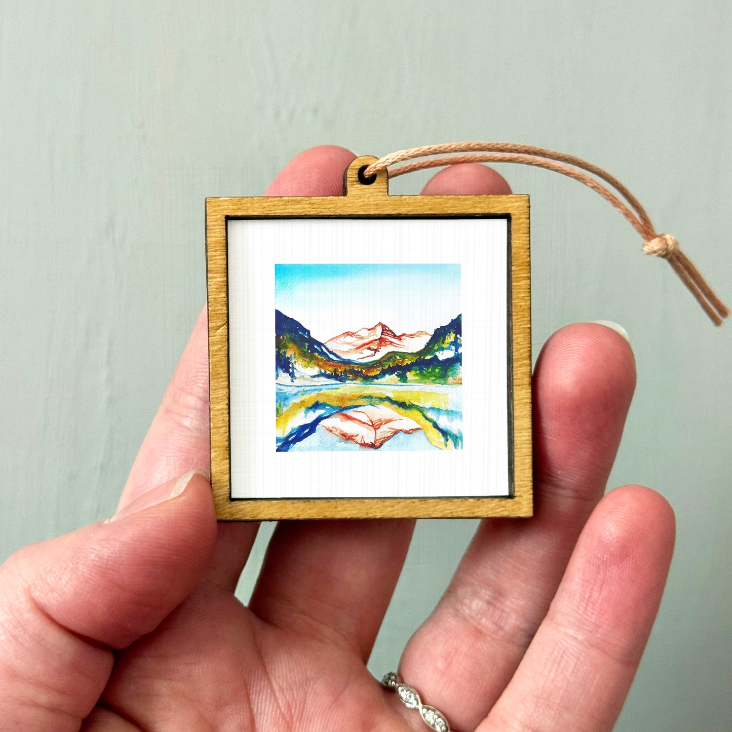 A hand holding a small wooden frame containing a watercolor painting of a mountain landscape.