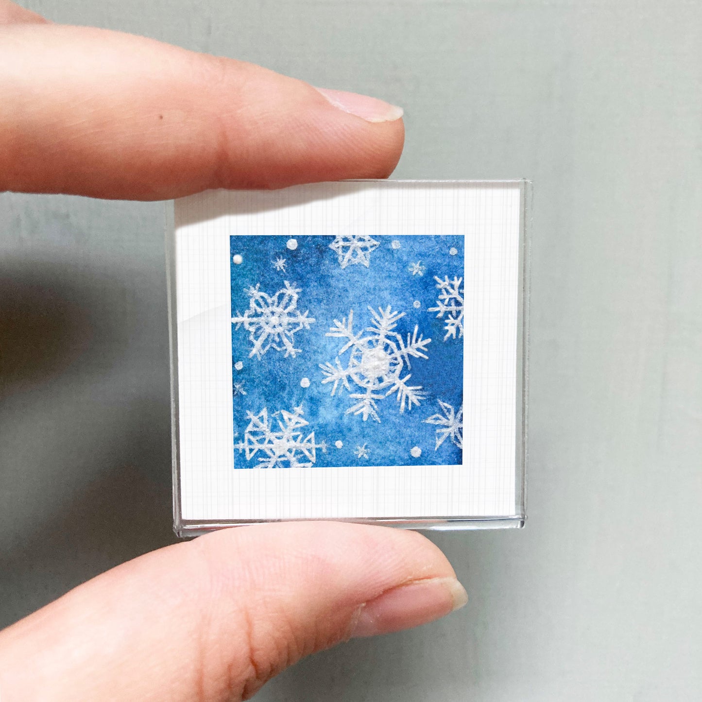 A hand is holding a small square picture with a blue background and white snowflakes.