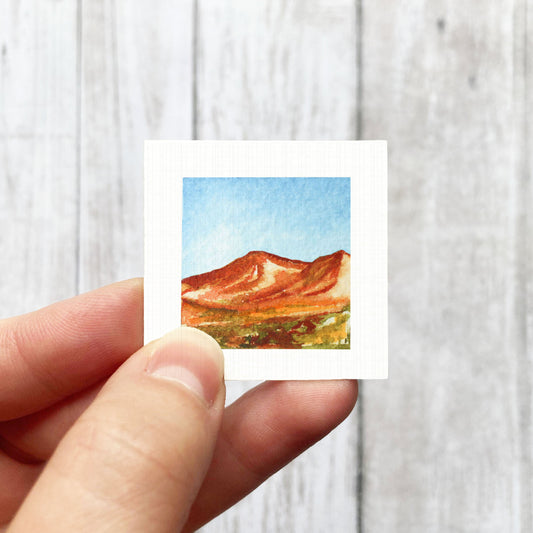A hand is holding a small square-shaped watercolor painting depicting a mountain landscape.