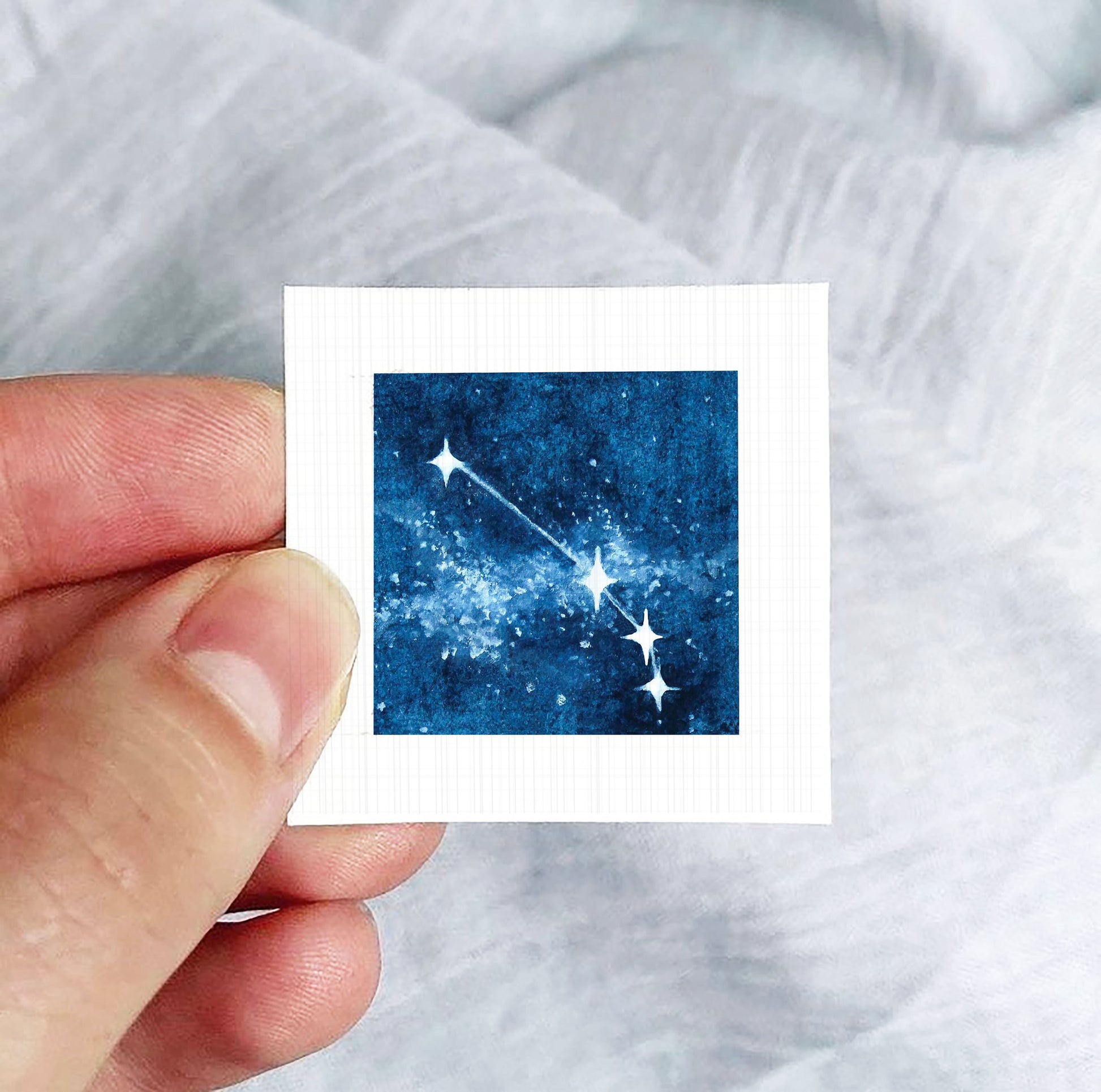 A hand is holding a small square-shaped object with a picture of a starry night sky on it.