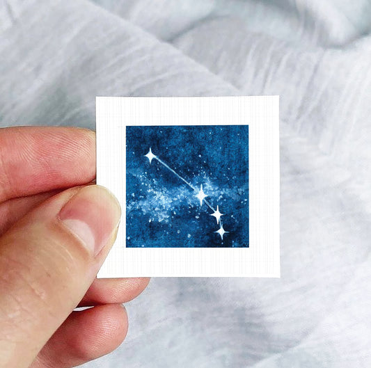 A hand is holding a small square-shaped object with a picture of a starry night sky on it.