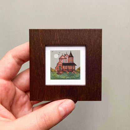 A hand is holding a small wooden frame containing a miniature painting of a house with a castle-like structure.