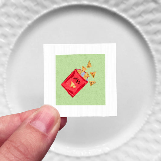 A hand is holding a small square with a red bag of chips and several chips spilling out onto a white plate.