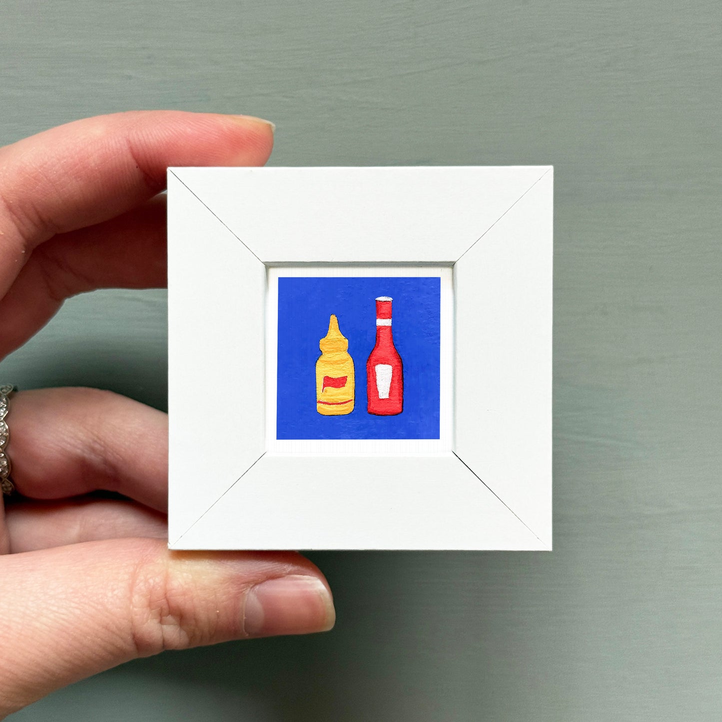 A hand is holding a small white square frame containing an illustration of two bottles of condiments, one yellow and one red.