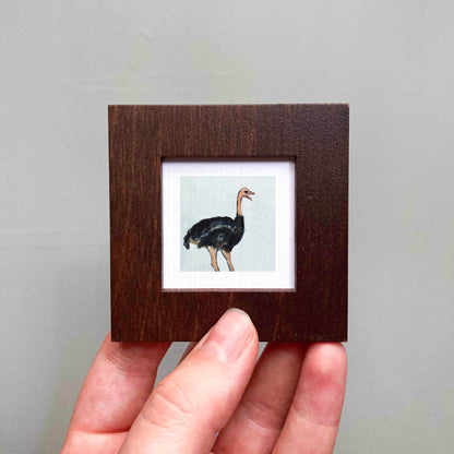 A person's hand is holding a small wooden frame containing a framed image of an ostrich.