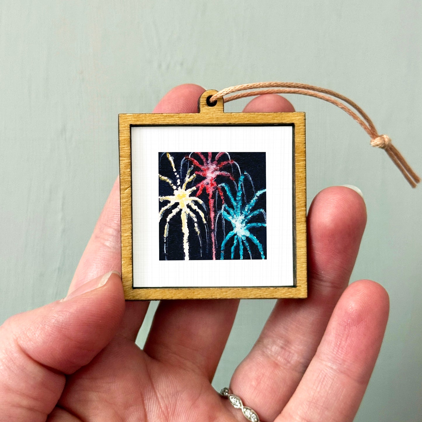 A hand holding a small wooden frame containing a picture of colorful fireworks.