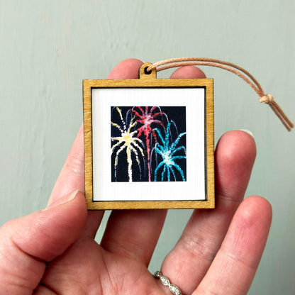 A hand holding a small wooden frame containing a picture of colorful fireworks.