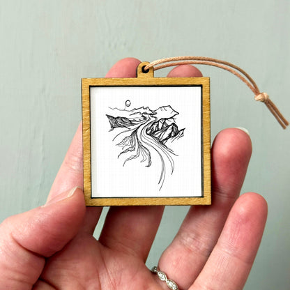 A hand holding a small wooden frame containing a black and white illustration of mountains and a winding river.
