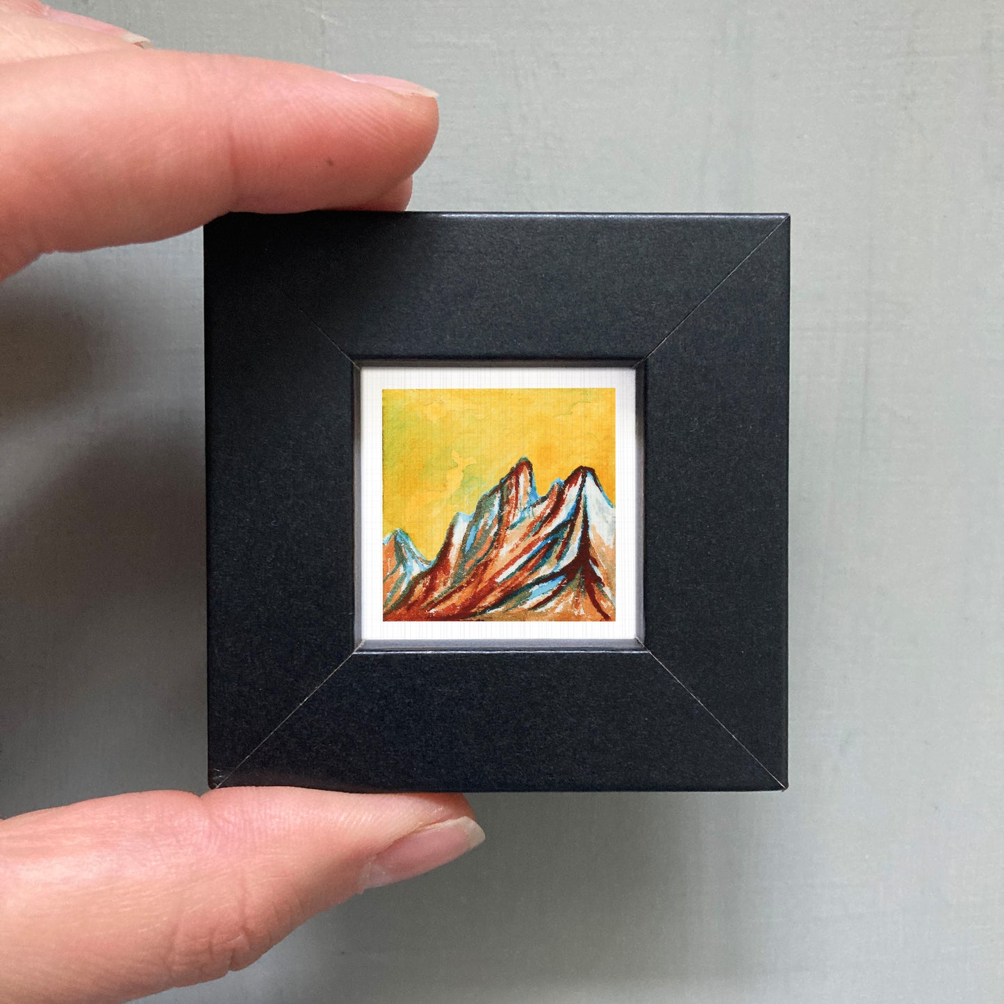 A person's hand holding a small black frame containing a colorful painting of mountains.