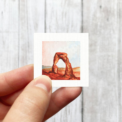 A hand holding a small square-shaped painting depicting a desert landscape with a large arch formation.