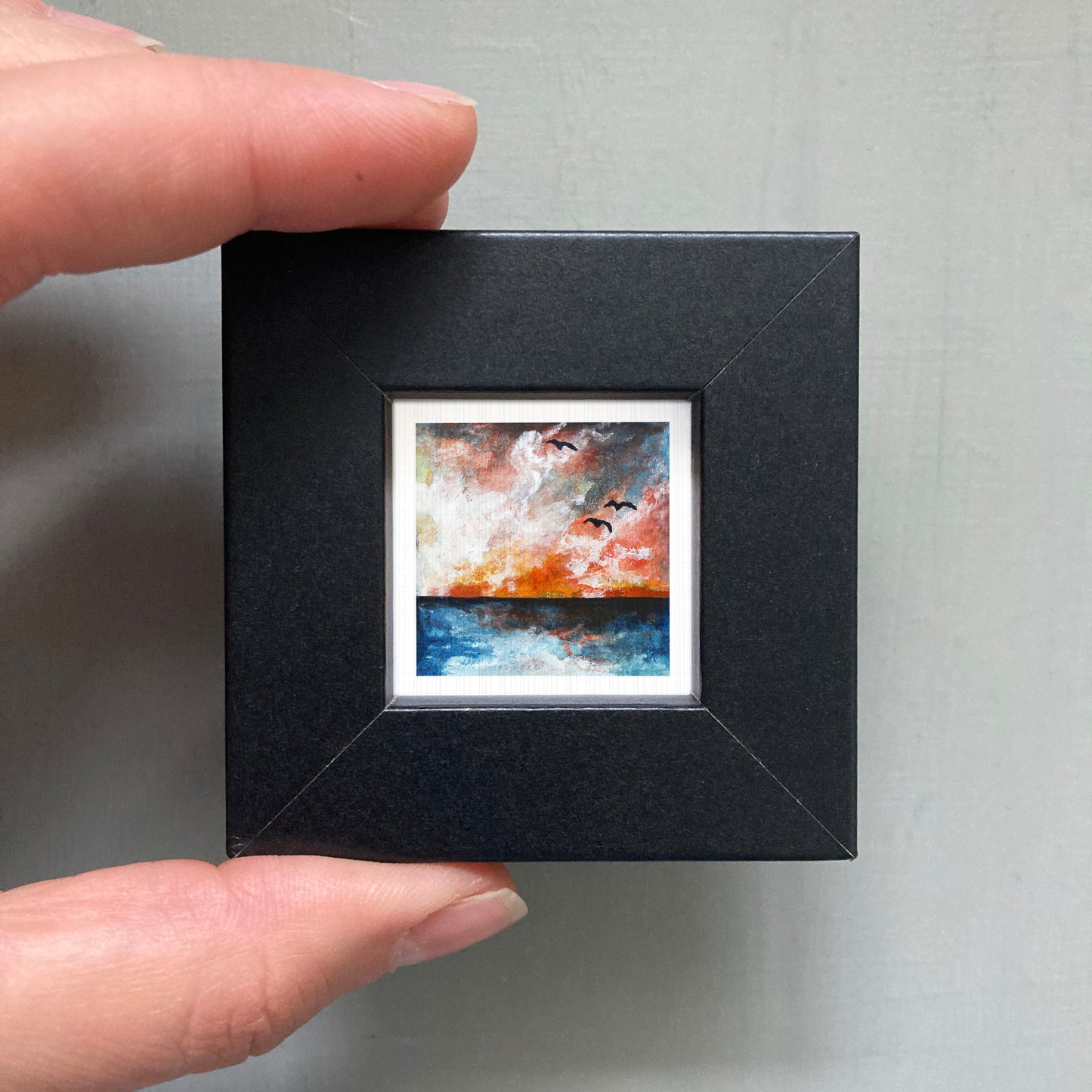 A person's hand is holding a small, square-shaped black frame containing a painting of a sunset over a body of water.