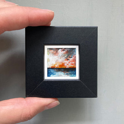 A person's hand is holding a small, square-shaped black frame containing a painting of a sunset over a body of water.