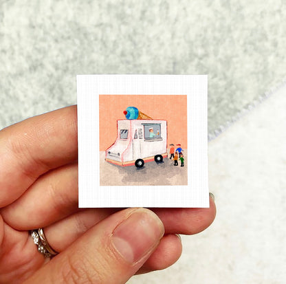 A hand holding a small square picture depicting an ice cream truck with people around it.