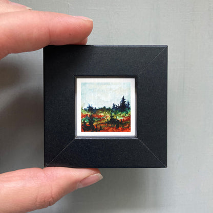 A person's hand holding a small, square-shaped black frame containing a miniature landscape painting.