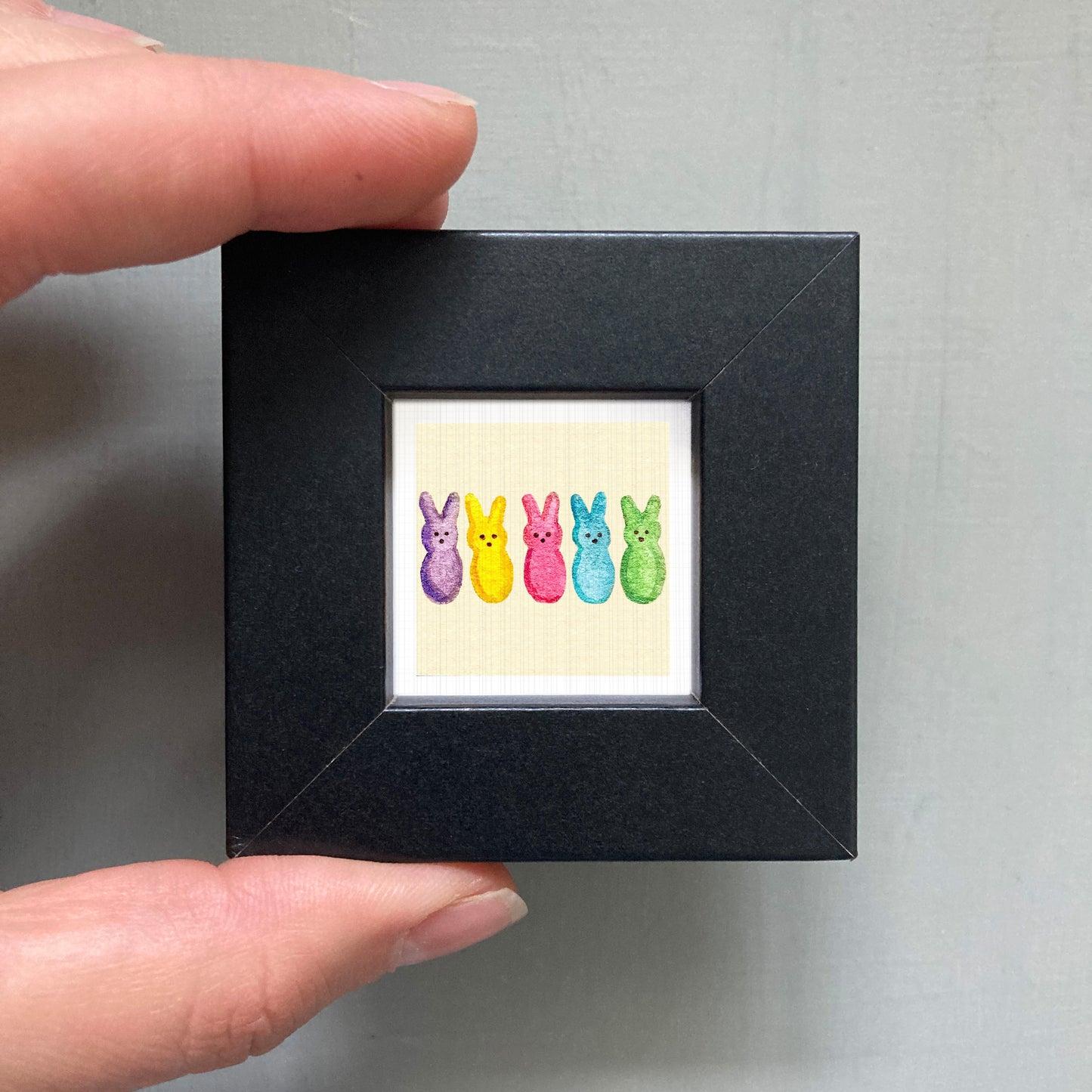 A hand is holding a small black frame containing a colorful illustration of five Easter bunny characters in various pastel shades.